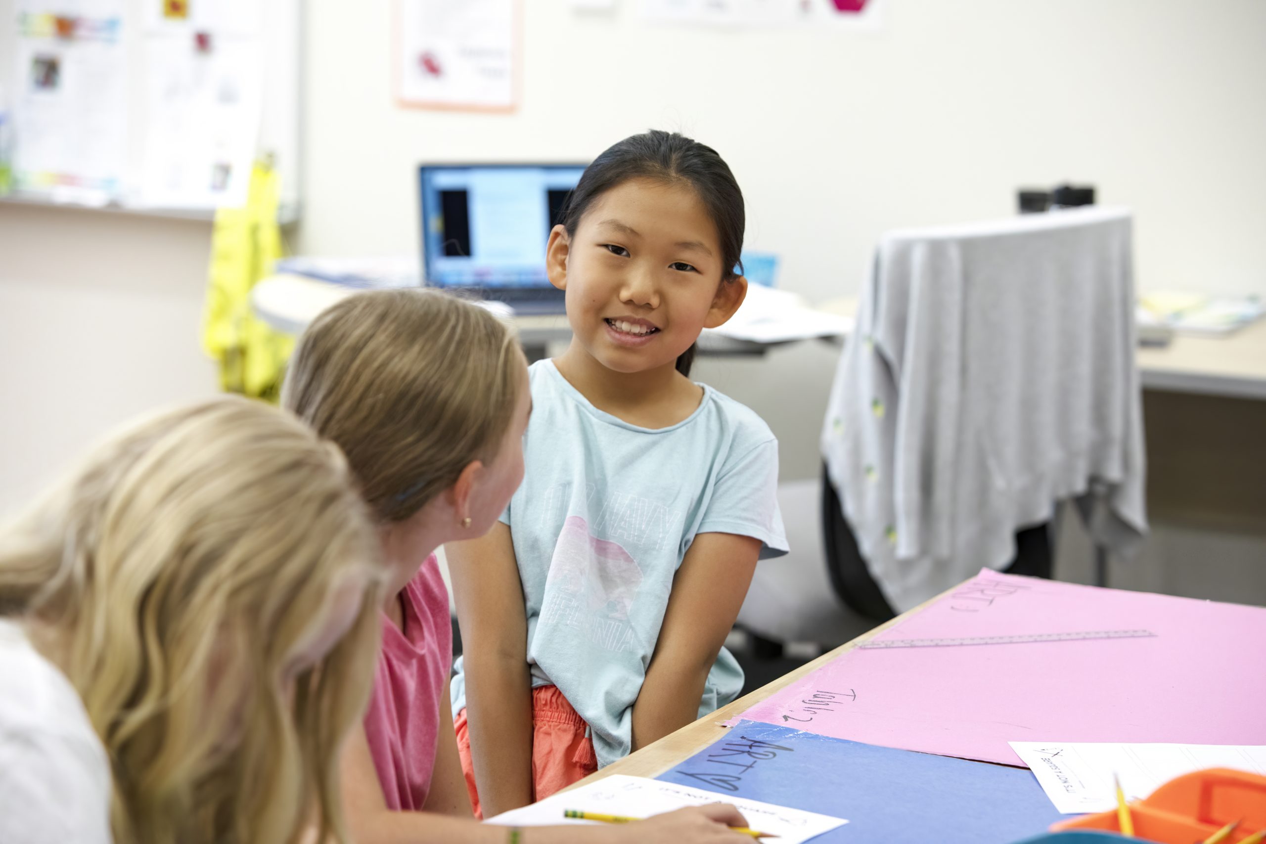 A young girl smiles at two classmates while working on a project at a classroom table with colorful papers.