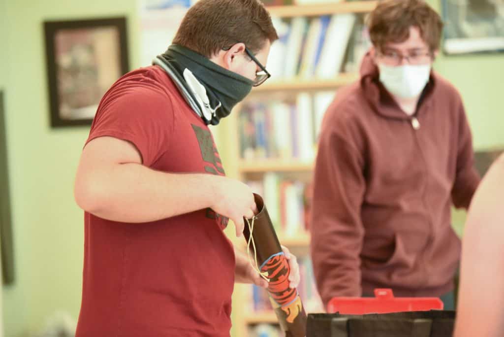 un estudiante de preparatoria con camisa roja y polaina facial carga un paracaídas en un cohete dentro de un aula