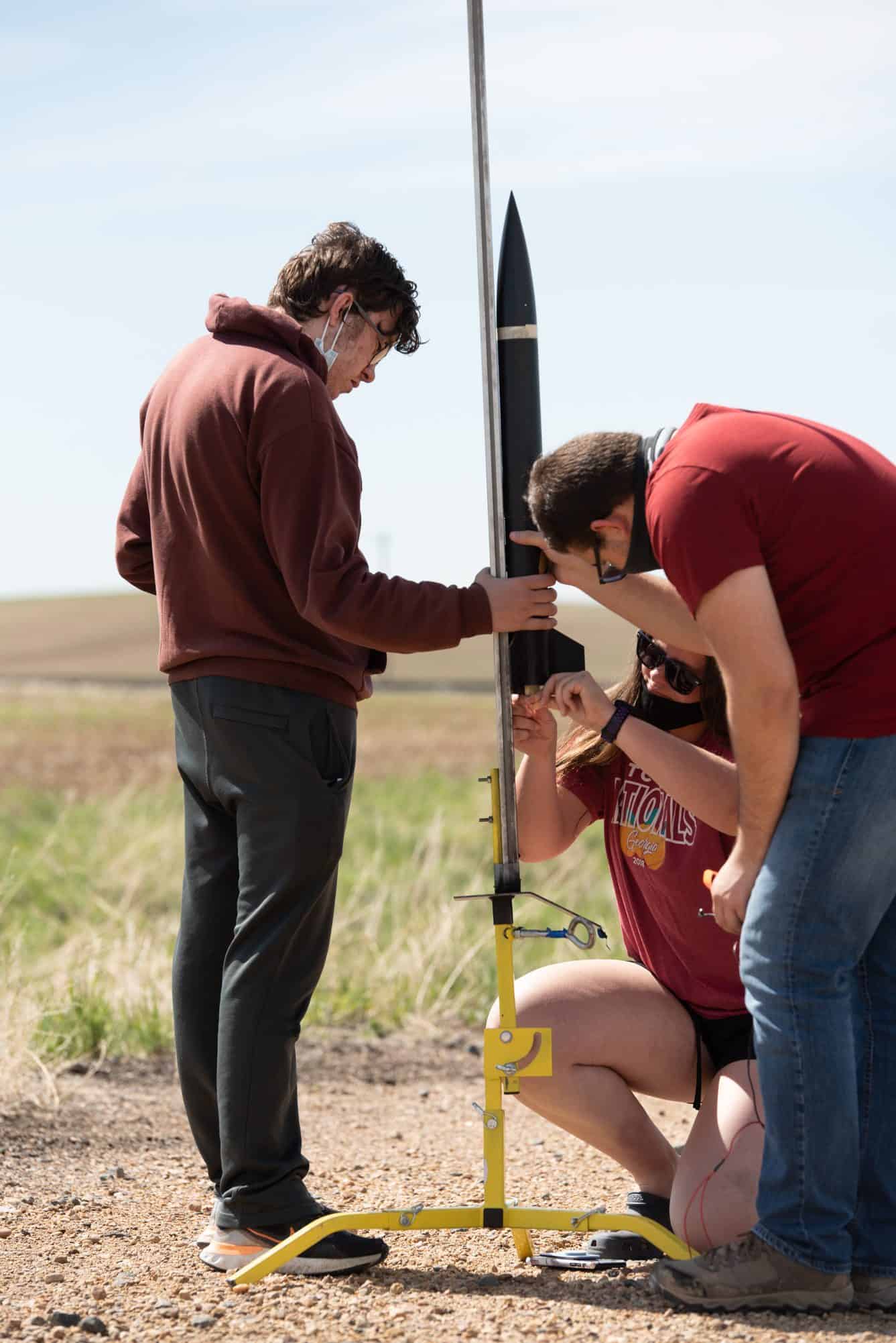 3 students gather and crouch around a rocket to prepare it to launch in a grassy field