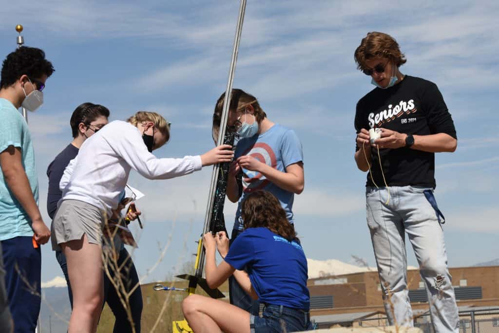 6 estudiantes de preparatoria -3 hombres y 3 mujeres- se reúnen alrededor de un poste inclinado en el que han montado un cohete que están a punto de lanzar.