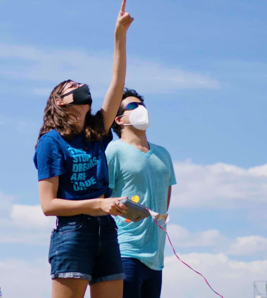 two high school students, one male and one female, in blue shirts look up outside on a bright day. The girl is pointing up.