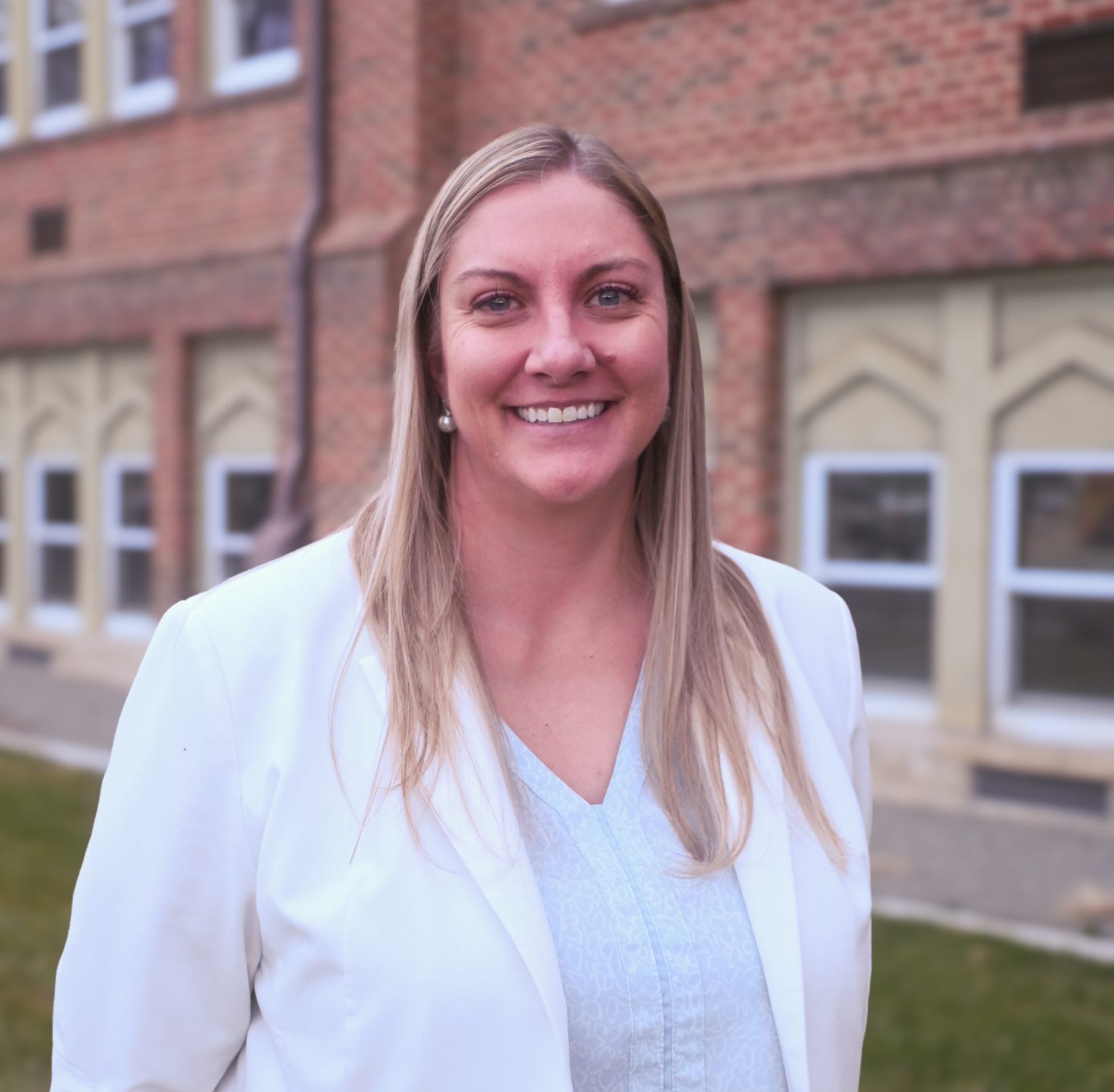 A smiling woman with long blonde hair in a white blazer stands in front of a brick building with large windows.