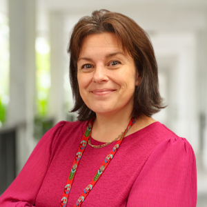 Mujer sonriente con una blusa rosa y un collar de colores, de pie en un espacio interior luminoso y moderno.