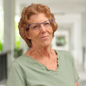 Una mujer mayor, sonriente, con el pelo rizado, gafas y una camiseta verde, está de pie en un pasillo luminoso.