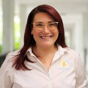 Una mujer pelirroja y con gafas sonríe en un pasillo luminoso; lleva una camisa blanca con el logotipo de «Nutrition Services».