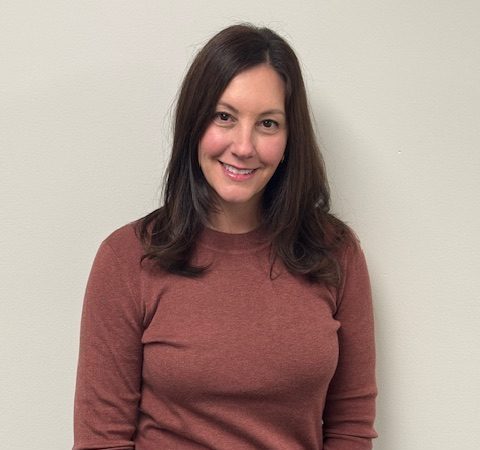 A woman with long brown hair smiles while standing against a light gray wall, wearing a brown sweater and black pants.