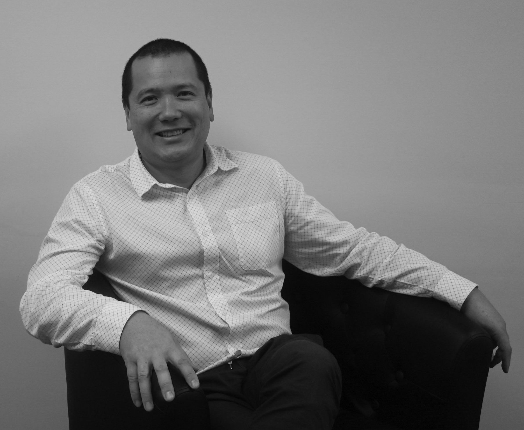 A smiling man in a checkered shirt sitting in a chair with a neutral background.