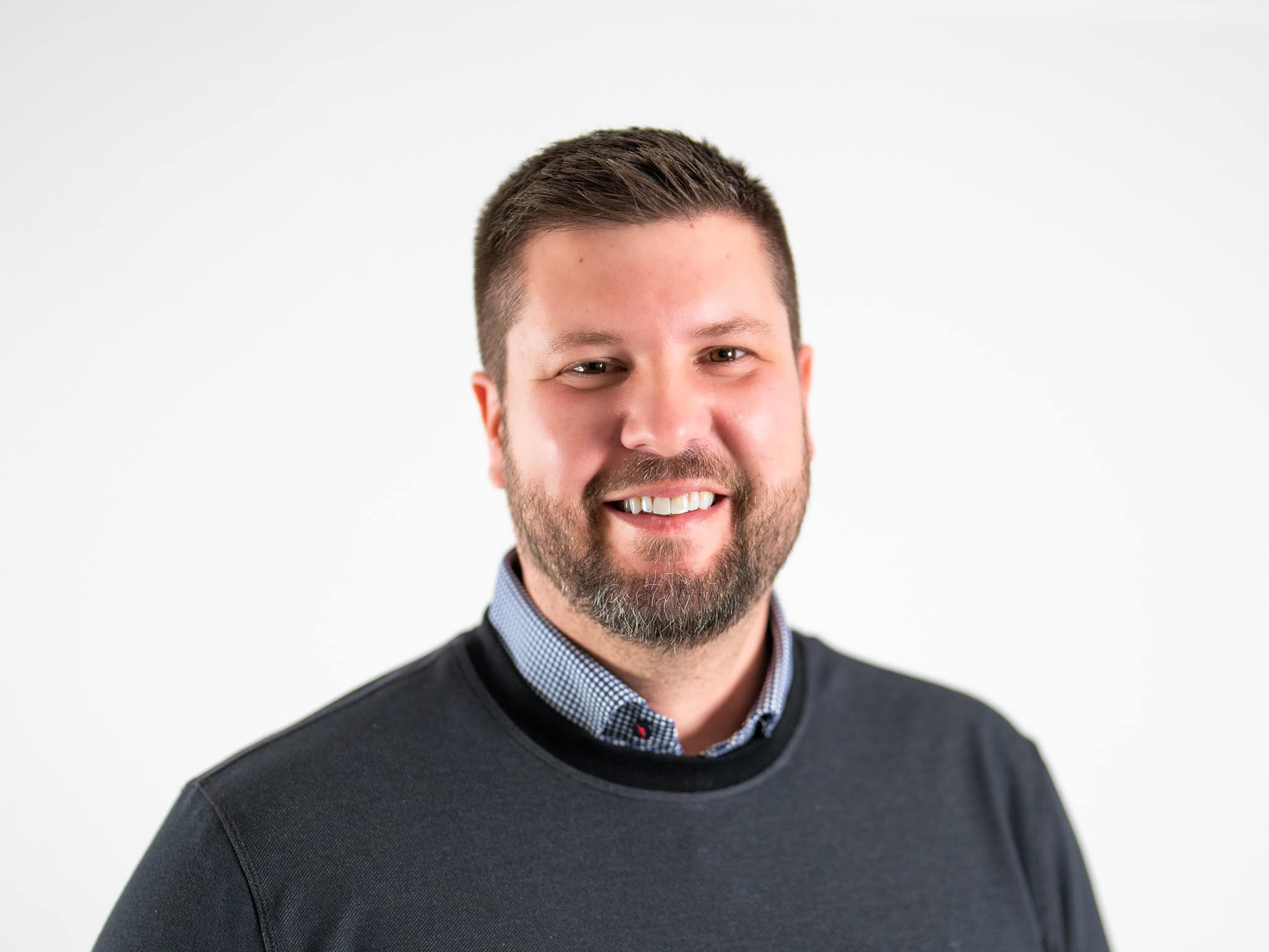 Smiling man with a beard, wearing a dark sweater over a checkered shirt, against a light background.