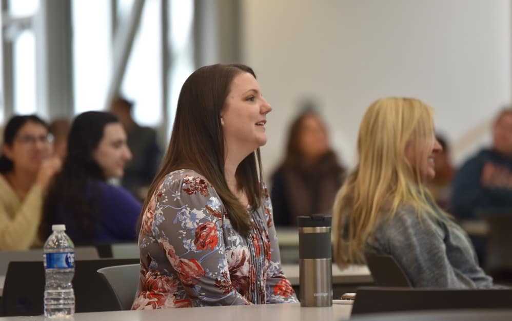 Profile view of a woman sitting at a table looking toward the speaker. Several other women are sitting at table next to and behind her. 