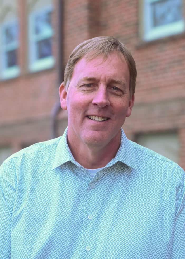 A smiling man with short hair wearing a light green patterned shirt stands outdoors in front of a brick building.