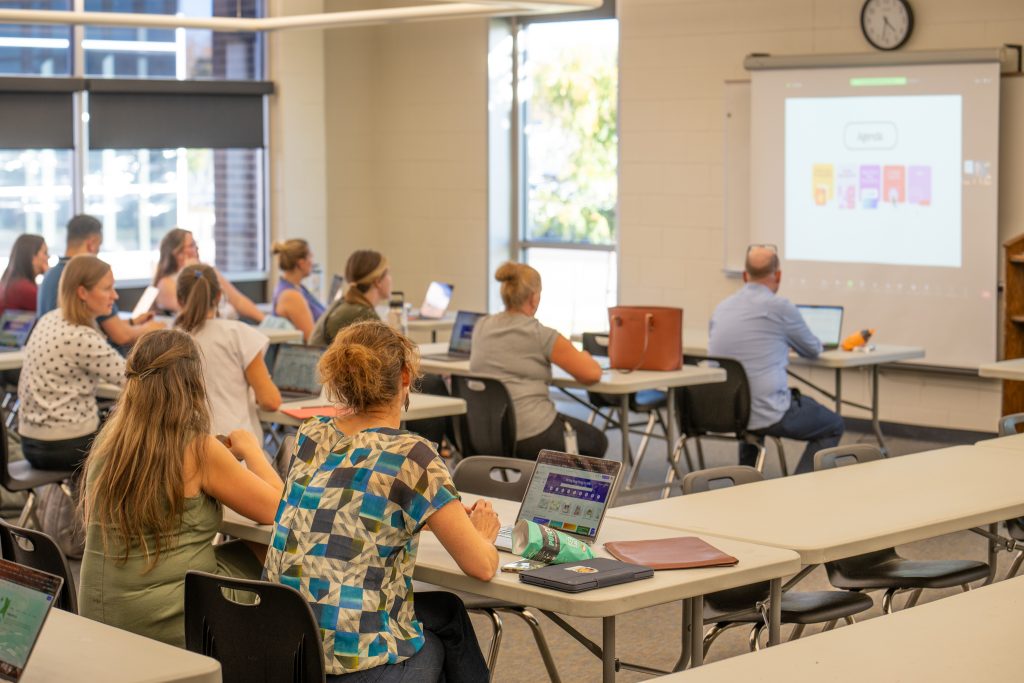 Group of male and female adult teachers sitting in a classroom watching an online presentation projected on a screen. 