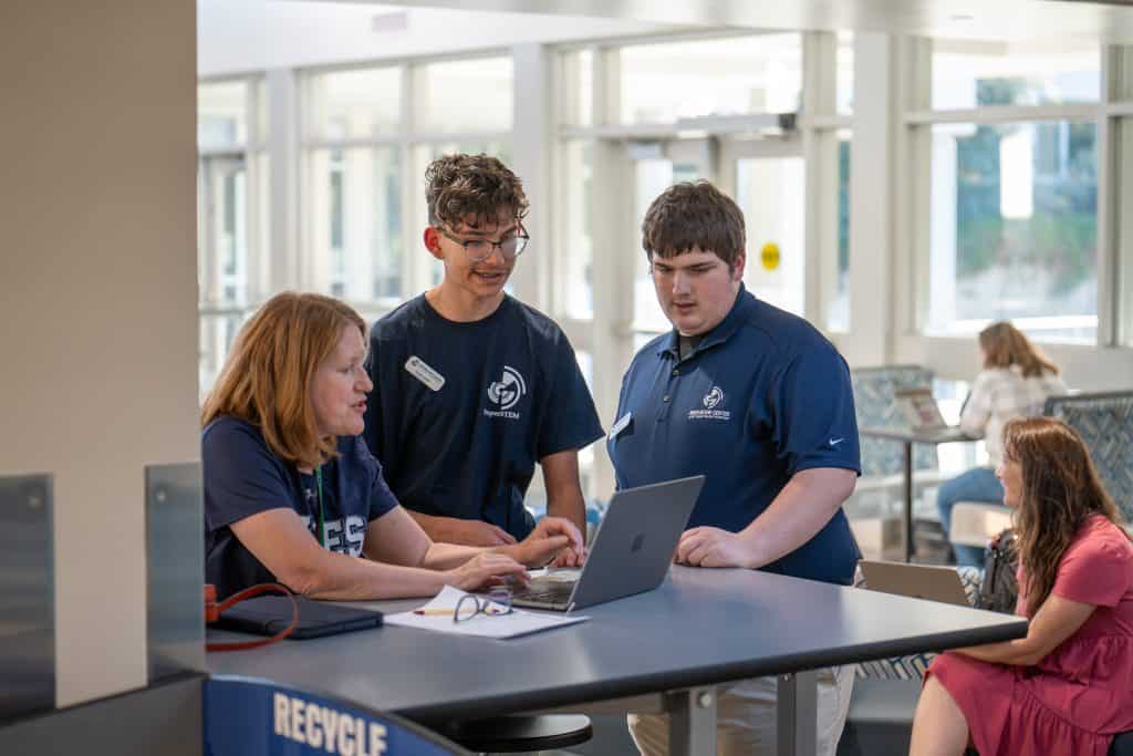 Woman and two high school students standing at a table working on a laptop. 
