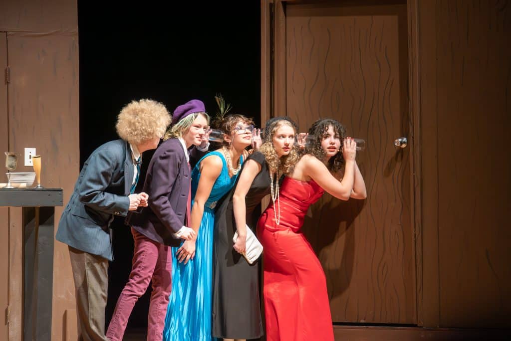 Six actors in Clue standing in a line. The first girl in line is holding a glass against the door, as if she's listening to what's on the other side. The others have a glass held against the person in front of them. 