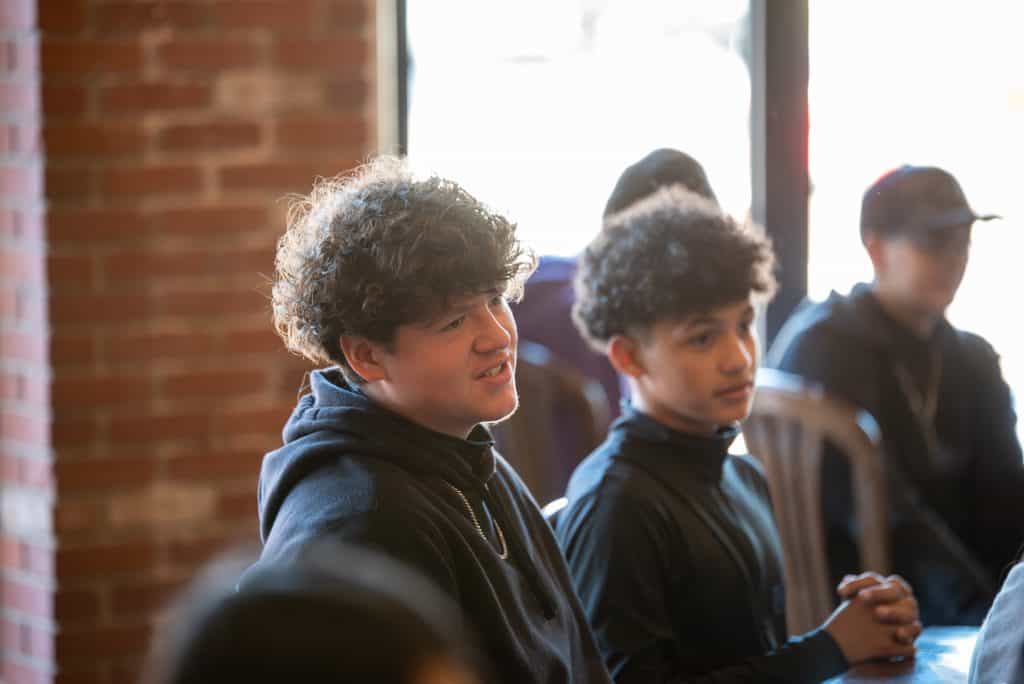 Dos preparatoria Hombres con cabello rizado escuchando a un orador en un restaurante local. Sentados en sillas con paredes de ladrillo y ventanas detrás.