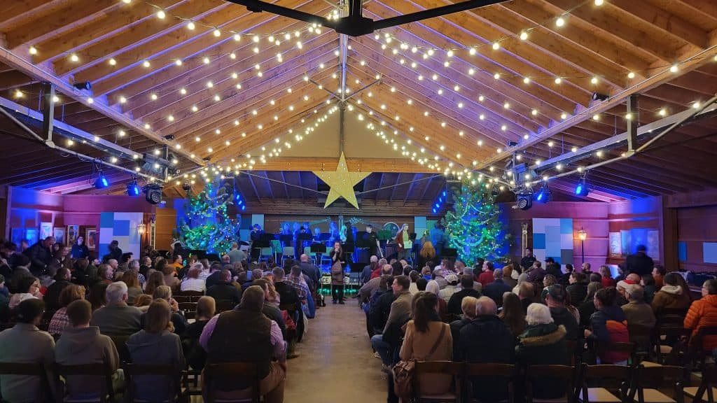 Vista desde el fondo de la sala mirando al escenario en Planet Bluegrass. Los miembros de la audiencia están sentados en sillas y una banda está en el escenario a punto de actuar. Hilos de luces Edison cuelgan en botines y filas del techo. Una estrella gigante cuelga del techo en el centro.  