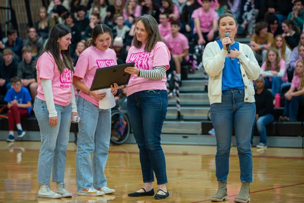 Two Mead High females and a female teacher are standing in the gym with a crowd of students blurred behind them. The teacher is holding a plaque that was given to her by Make-a-Wish. She is showing it to the female students next to her. A female from Make-a-Wish is speaking on a microphone to the right of them. 