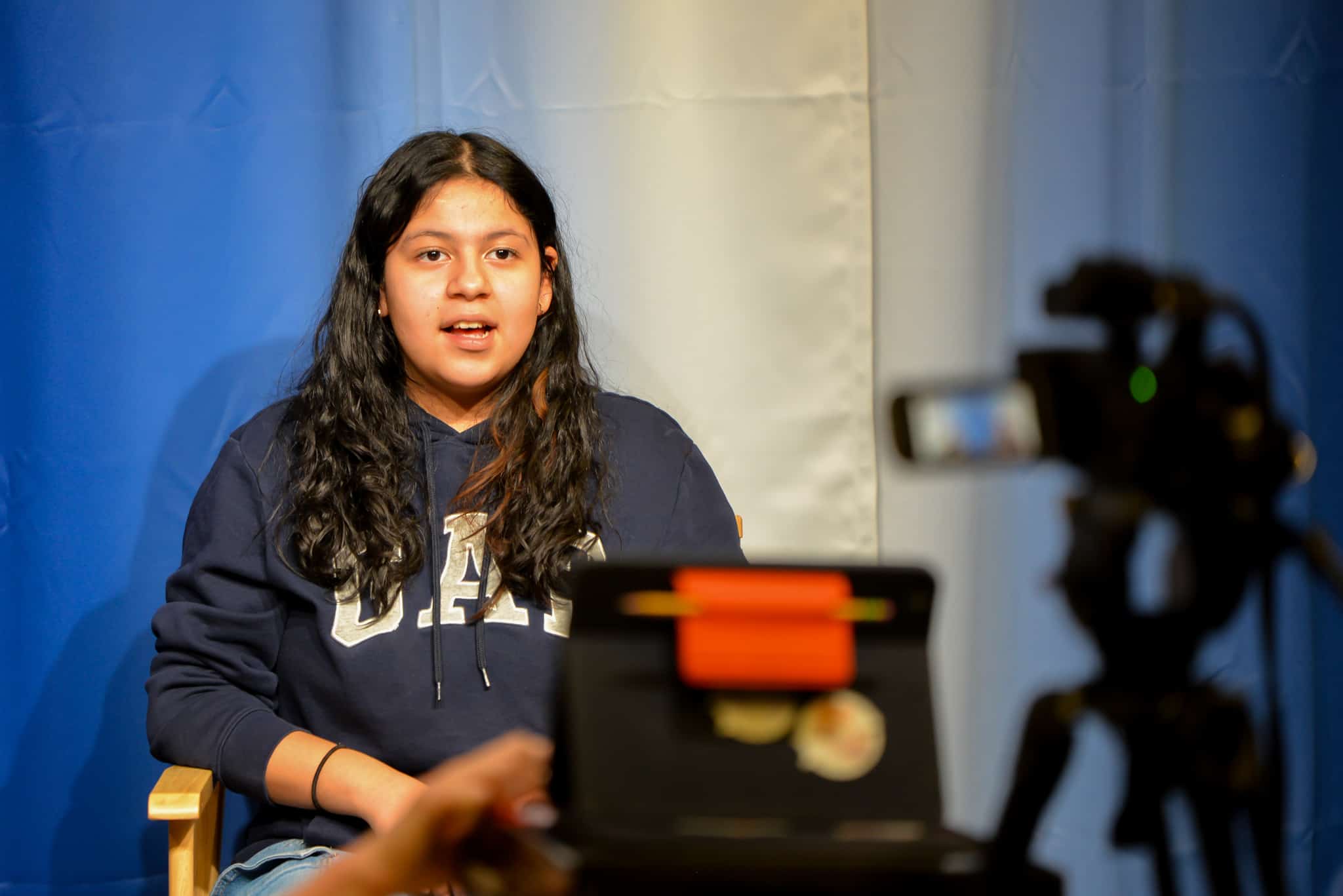 Hembra secundaria estudiante sentado en una silla frente a una cámara. Ella está hablando en cámara durante un noticiero que está siendo grabado.