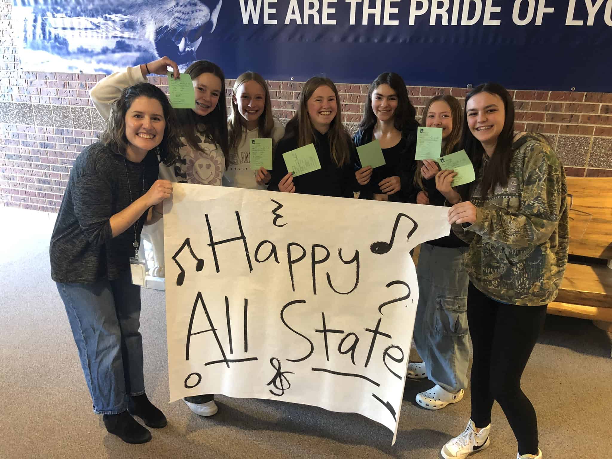 Lyons Middle School Choir teacher on the left with six female choir students to the right of her holding cards that say there were selected for all state choir. They are holding a banner that says Happy All State.