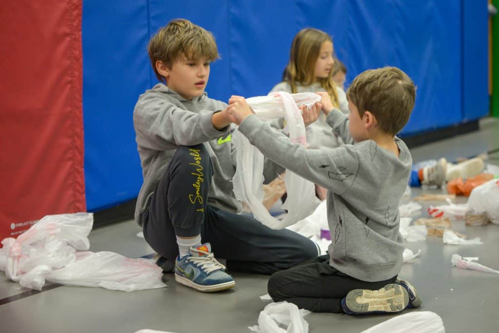  Un alumno de 5º curso y otro de 1º trabajando en un proyecto de manualidades haciendo cuerdas para saltar con bolsas de plástico. Están sentados en el suelo uno frente al otro y tienen los brazos extendidos mientras se agarran a las bolsas. 