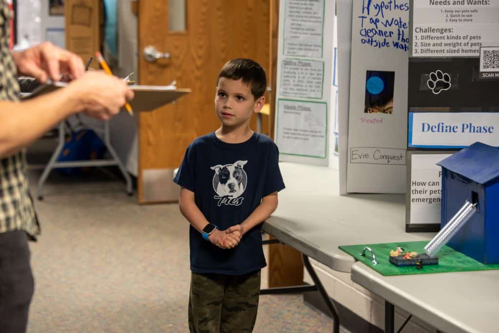 Third grade boy, Caleb, standing in front of the table his STEAM project is sitting on. The model of the inflatable emergency pet slide he created is on the table next to him. He is holding his hands in front of him and looking up slightly with his eyes at the judge in front of him.