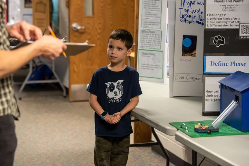 Caleb, un niño de tercer curso, de pie delante de la mesa en la que está su proyecto STEAM . La maqueta del tobogán hinchable de emergencia para mascotas que ha creado está sobre la mesa a su lado. Tiene las manos delante y mira ligeramente hacia arriba con los ojos puestos en el juez que tiene delante.