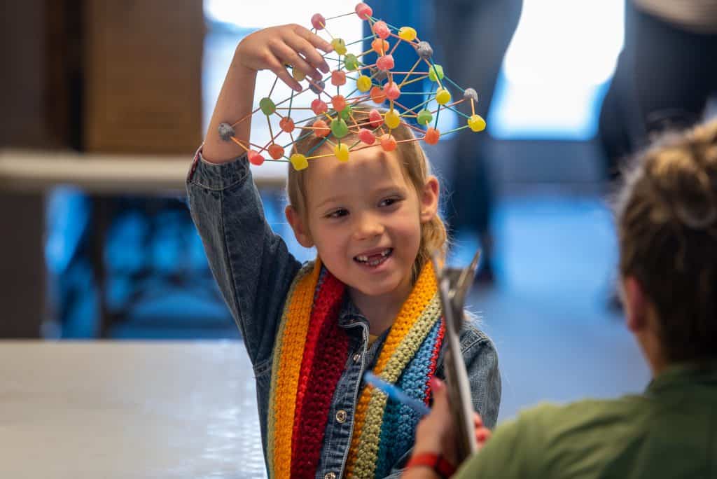 Niña de tercer curso con una colorida bufanda de punto. Sonríe mientras sostiene parte de su proyecto de ciencias, una molécula hecha de gominolas y palillos. 