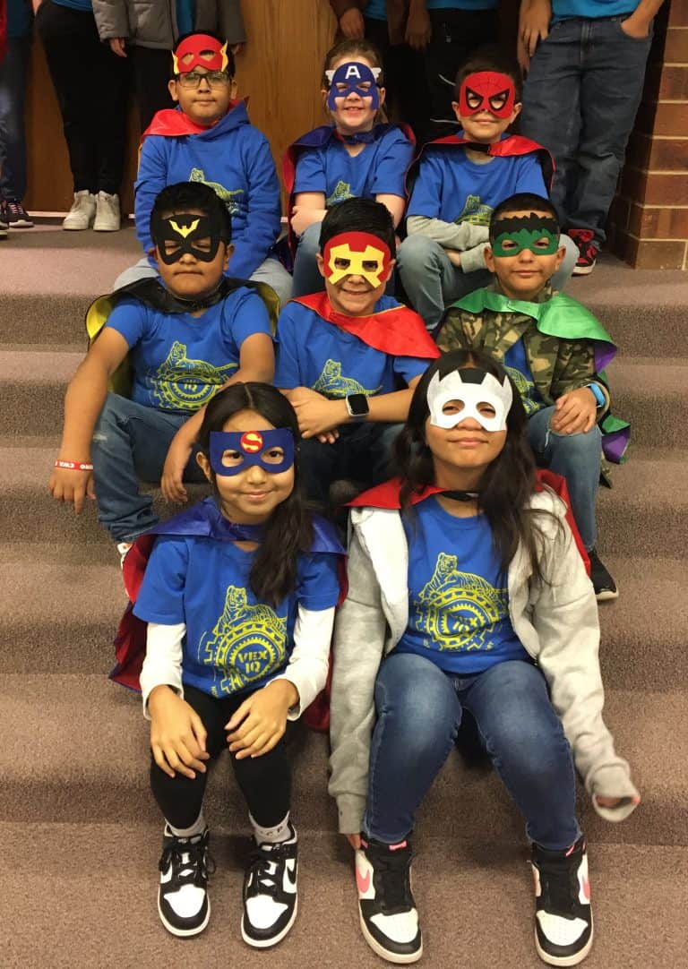 Eight elementary students from Northridge sitting on carpeted steps in three rows. The back and middle rows have three bows across and the front row has two girls. They are wearing superhero masks over their eyes along with capes for the superhero-themed robotics tournament.