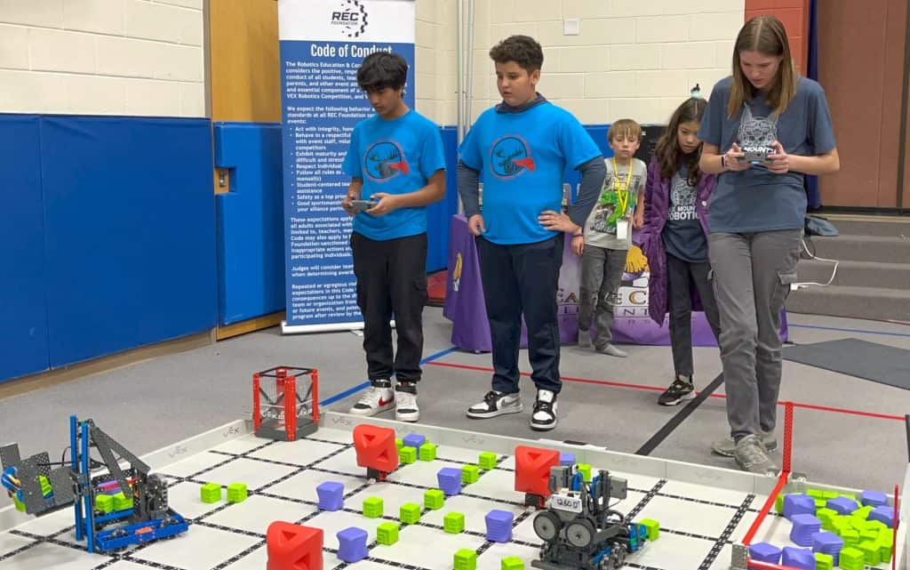 Dos estudiantes varones de 5º curso de Indian Peaks de pie, uno al lado del otro, delante de un campo de robótica. El chico de la izquierda sostiene un mando y mueve su robot por el campo. Una estudiante más alta de otra escuela está a la derecha de ellos y también controla su robot con un mando.