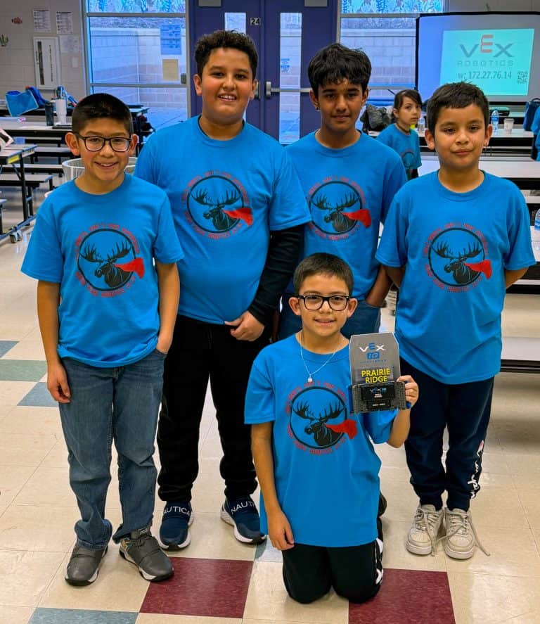 A group of five elementary school boys, four standing behind one stuent who is on their knees in front holding a trophy for robotics. They're wearing matching t-shirts with and Indian Peaks Elementary Robotics log on it. They're standing in a school.