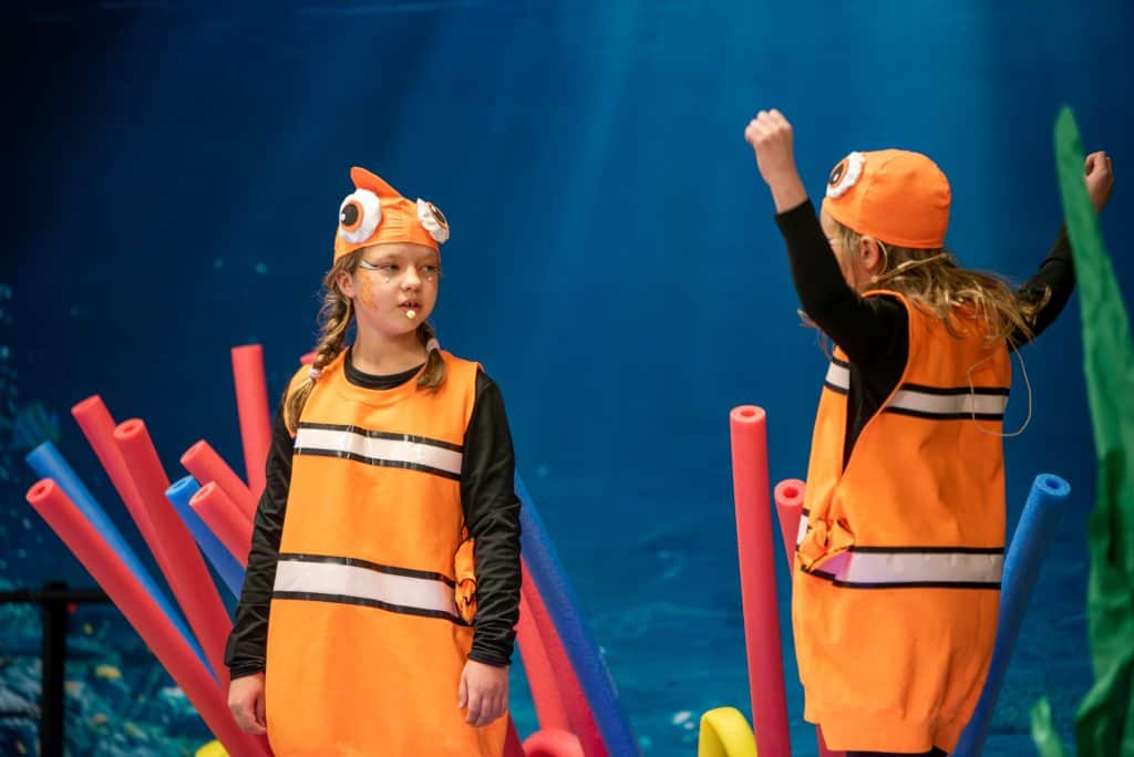 Dos estudiantes de primaria en un escenario vestidas de peces payaso. Están frente a frente. El telón de fondo hace que parezca que están bajo el agua. 