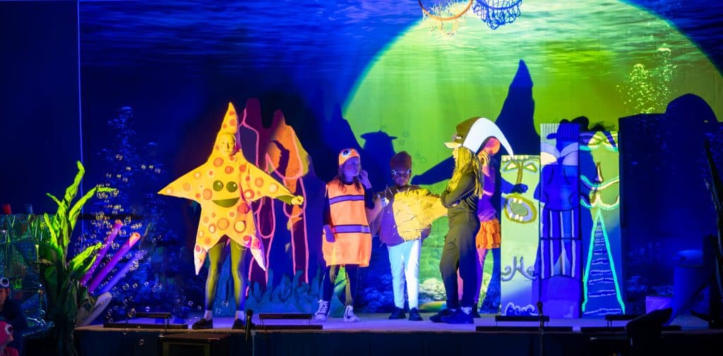 Five elementary students dressed as starfish and fish standing in a scattered line on a stage made to look like they're underwater. 
