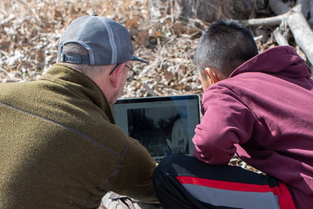 Mirando desde atrás, un profesor varón a la izquierda y un alumno varón de secundaria a la derecha están sentados en el suelo en una zona de espacio abierto mirando las fotos de la tarjeta SD que sacaron de su cámara de rastreo. En la pantalla del portátil se ve un ciervo.