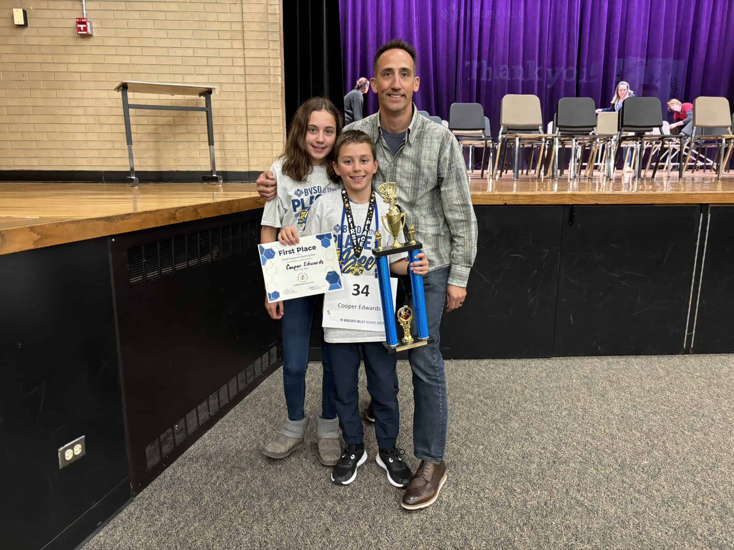 Cooper's 7th grade sister on the left, Cooper in the middle front, and Cooper's dad on the right. Standing and smiling at the camera. Cooper is holding an award and first place trophy for winning the regional spelling bee.