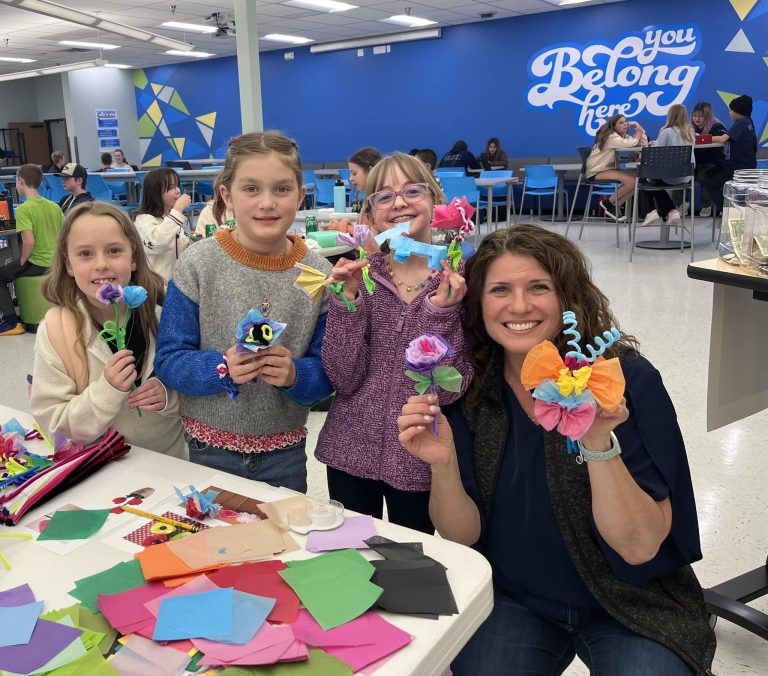 Three elementary students standing next to female principal on in front of a craft table and holding up pipe cleaner flowers they made.