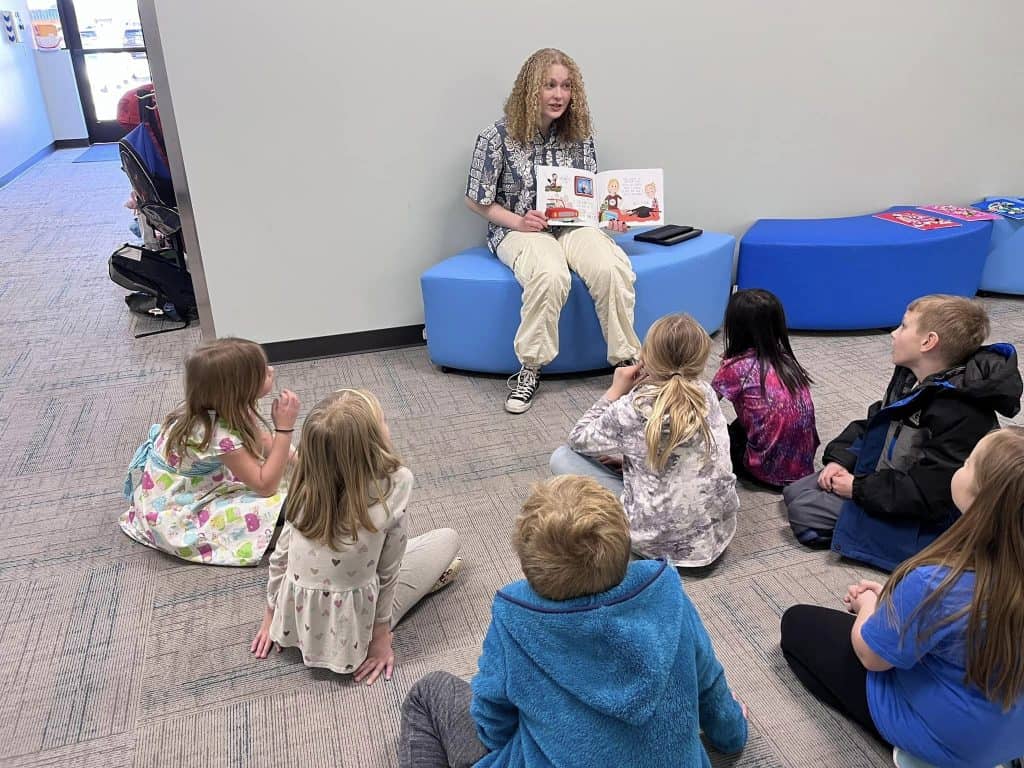 Female high school student sitting in front of elementary students and reading a book to them.