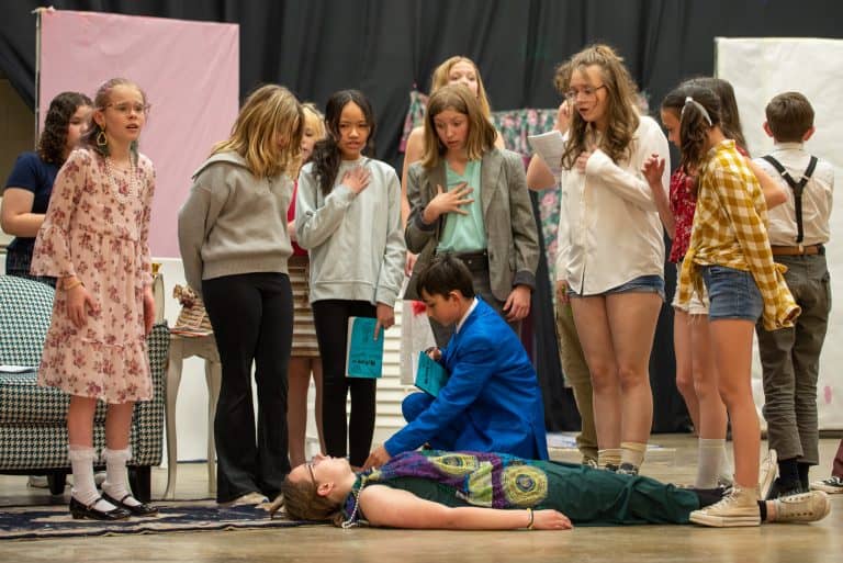 A line of female students standing above a male student who is checking for the pulse of a female student laying on the stage during a play.