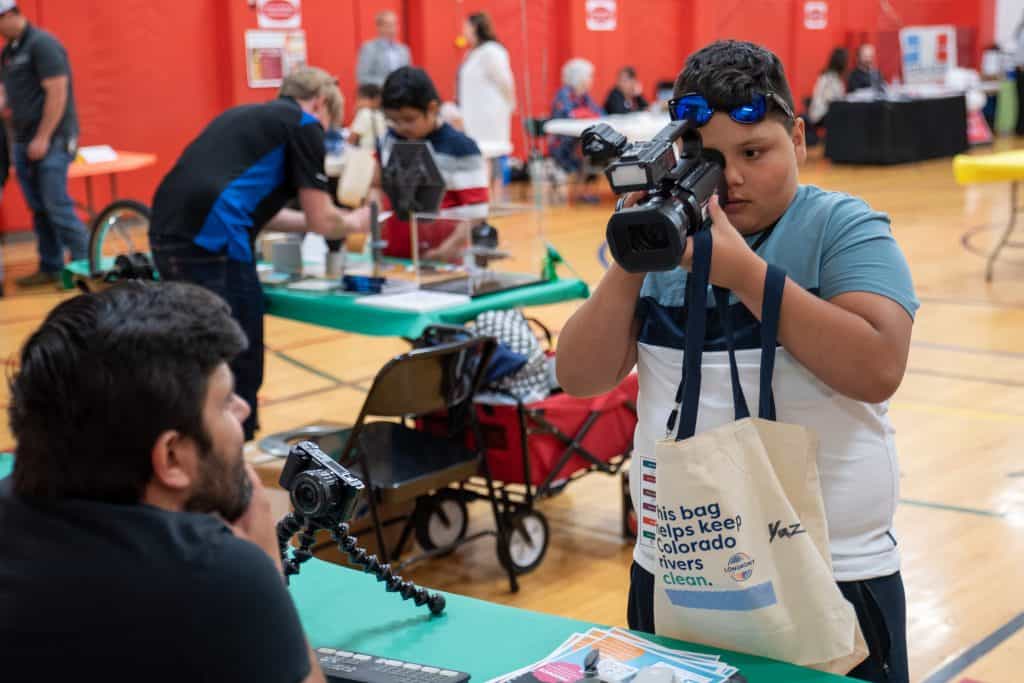 Adult male sitting at a table on the left while a male elementary student on the right looks through a video camera at Career Day.