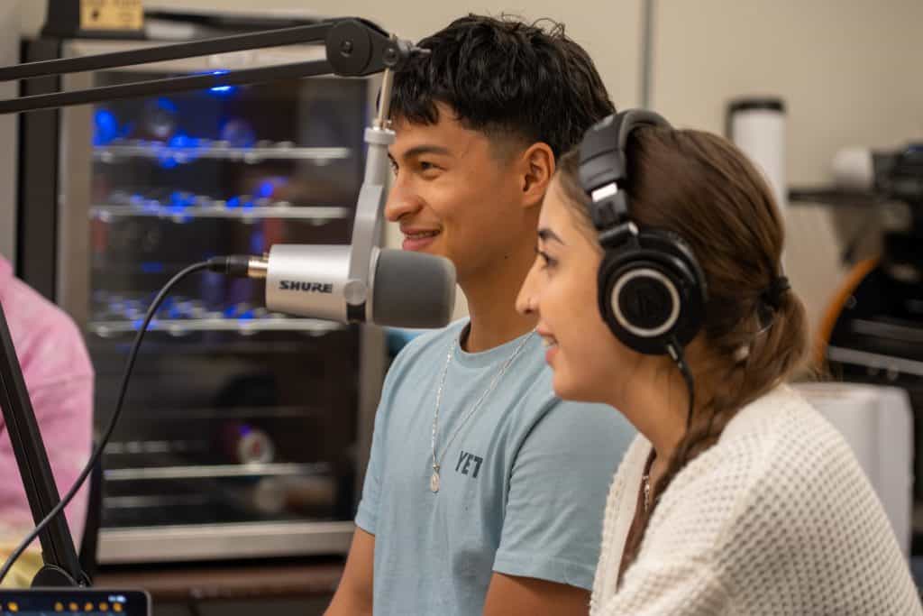 Male student in center of photo with female on the right. Both are sitting in front of a podcast microphone. 