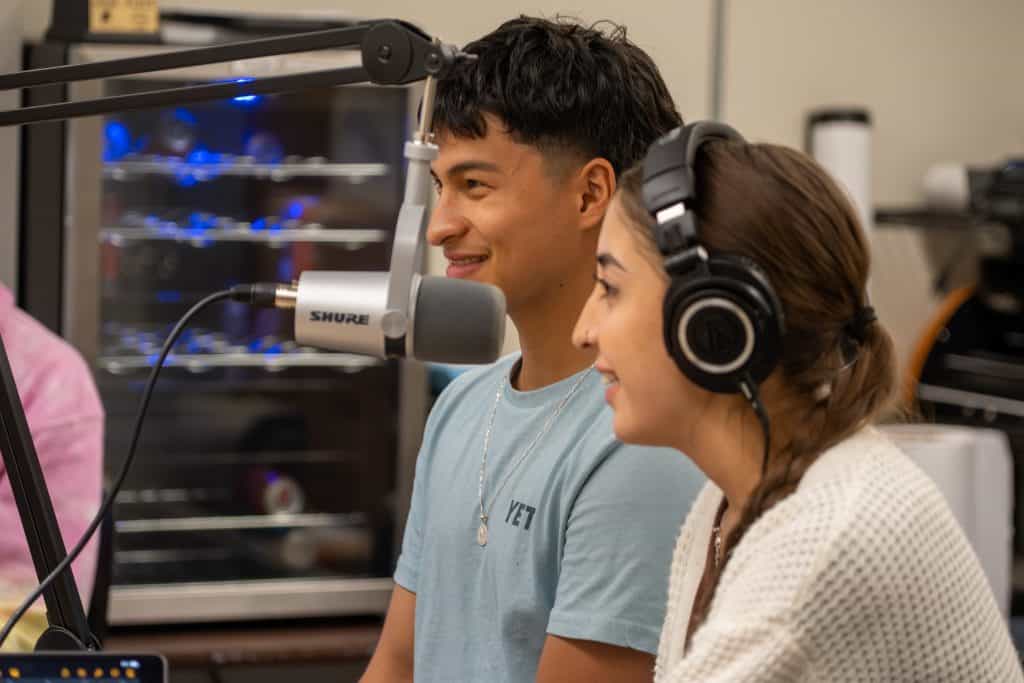 Estudiante masculino en el centro de la foto con una mujer a la derecha. Ambos están sentados frente a un micrófono de podcast. 