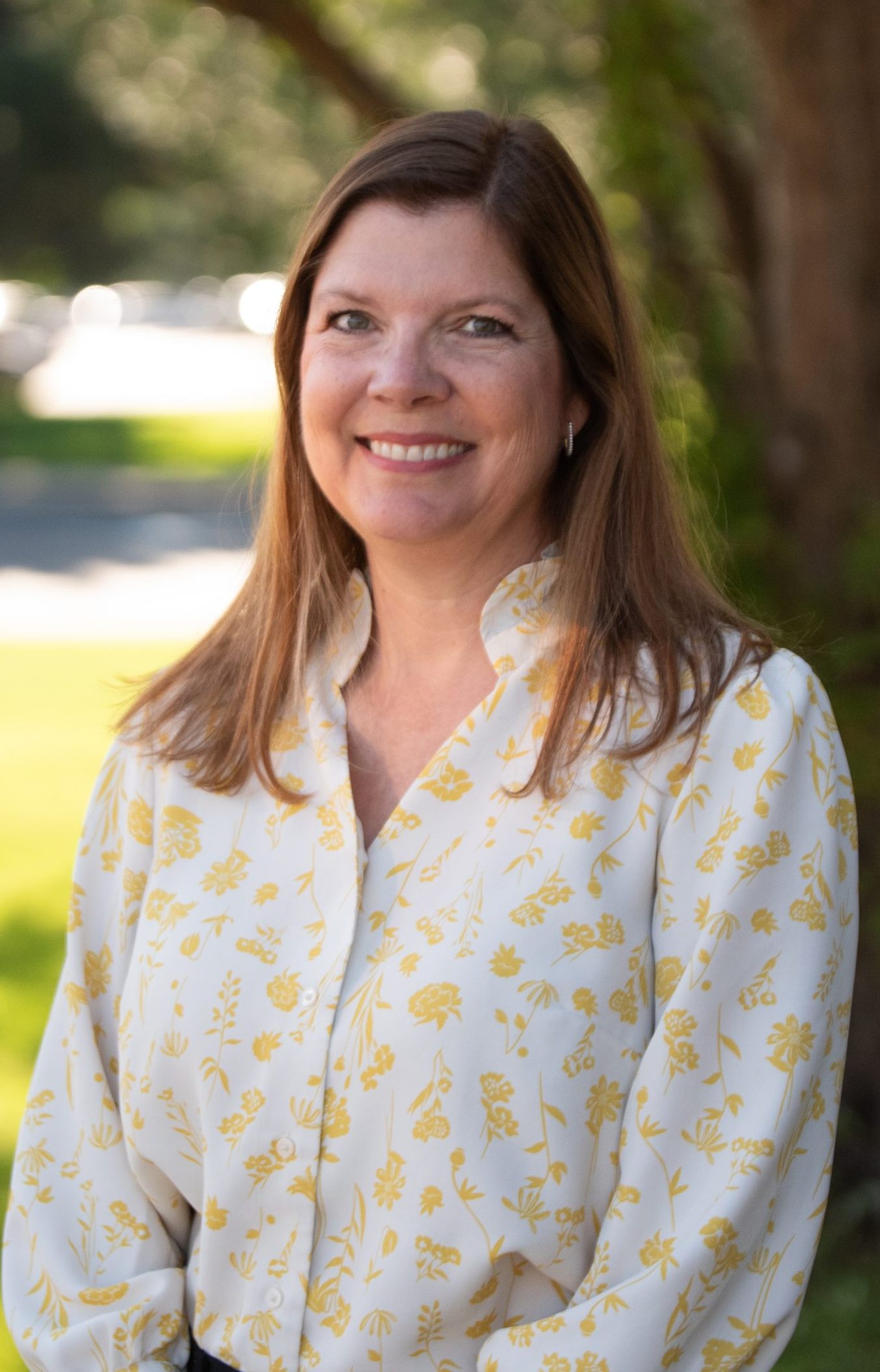 A smiling woman with long hair stands outdoors, wearing a white shirt with yellow floral patterns and black pants.