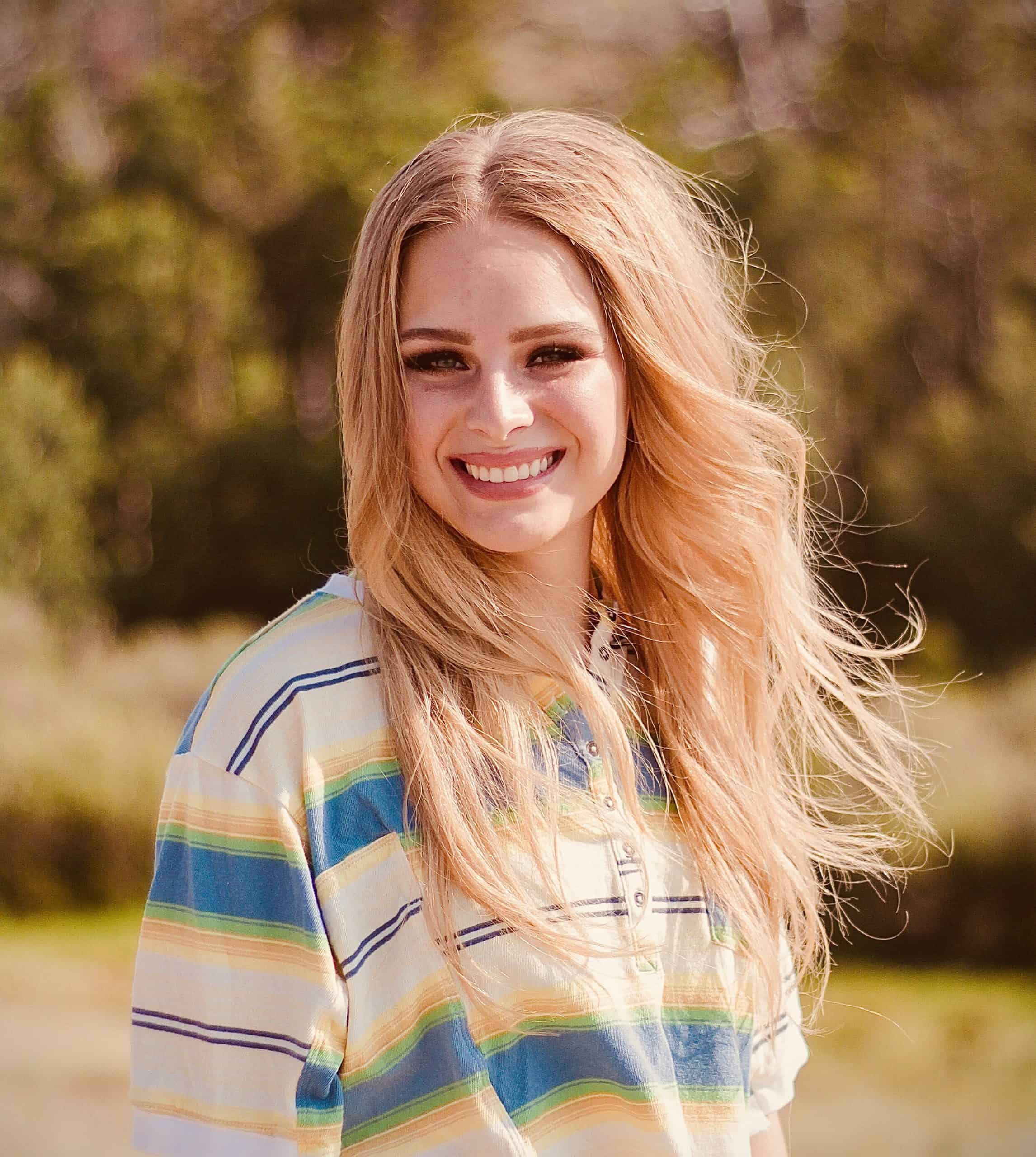 A smiling young woman with long, wavy blonde hair stands outdoors, wearing a colorful striped shirt.