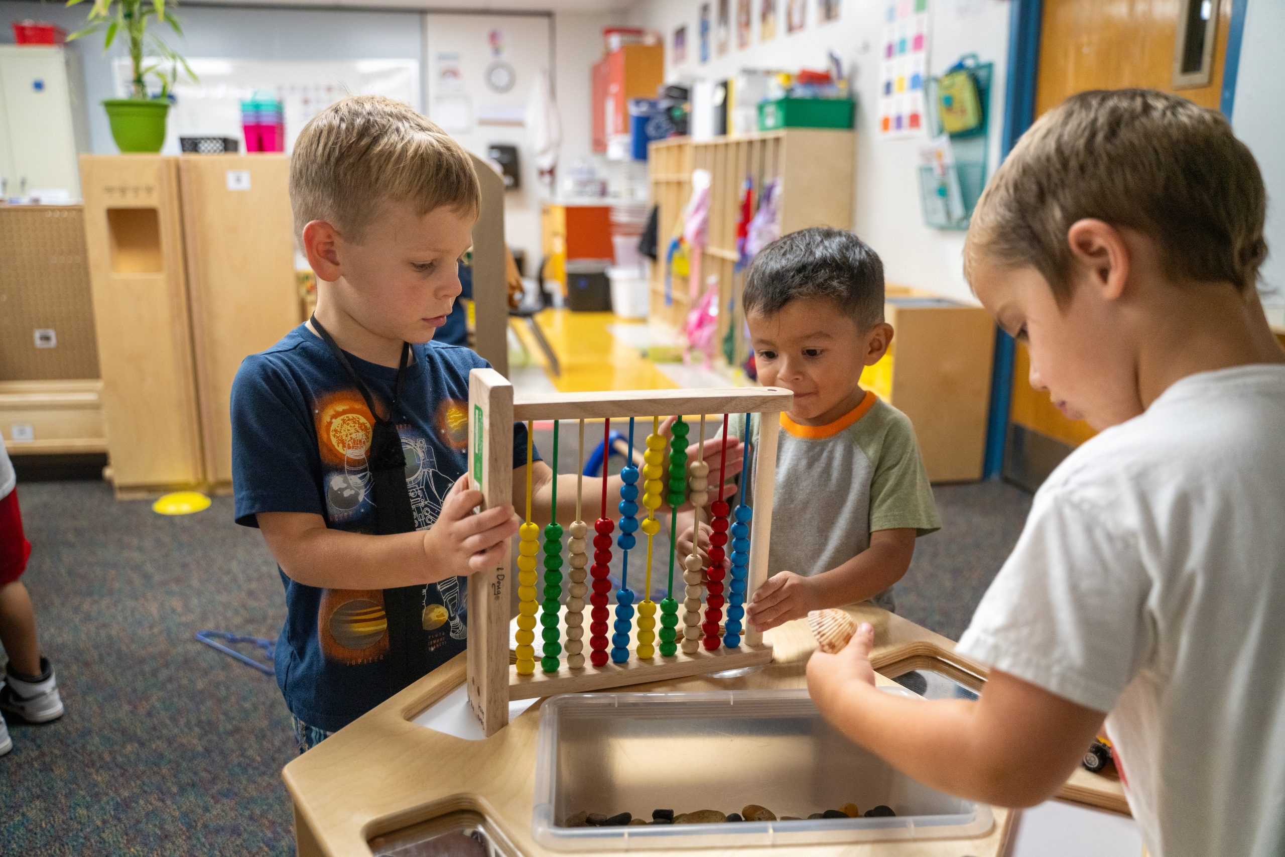 Three young children are playing with a colorful abacus and exploring small objects in a classroom setting.