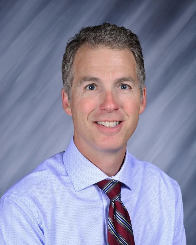 Smiling man with short, light brown hair, wearing a light purple shirt and a striped tie, against a gray backdrop.
