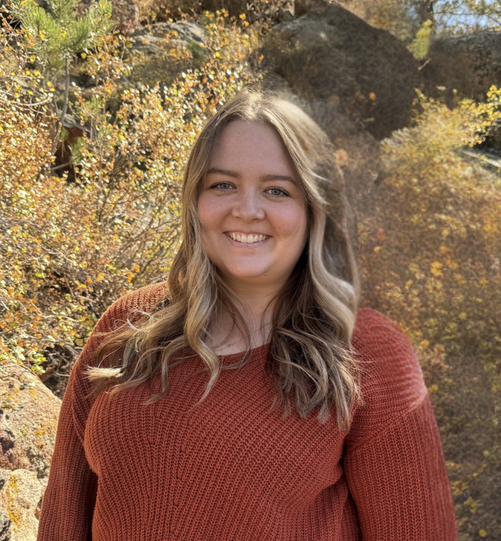 A smiling woman with long hair wears an orange sweater, standing outdoors among autumn foliage and rocky terrain.