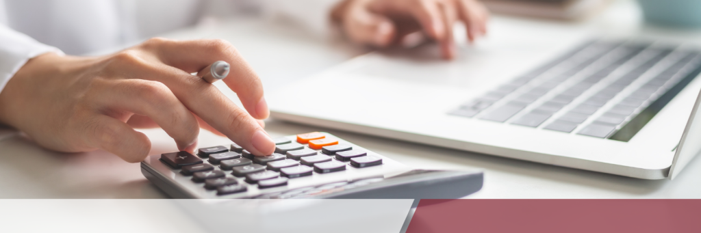 A person uses a calculator with one hand while typing on a laptop with the other, on a desk.
