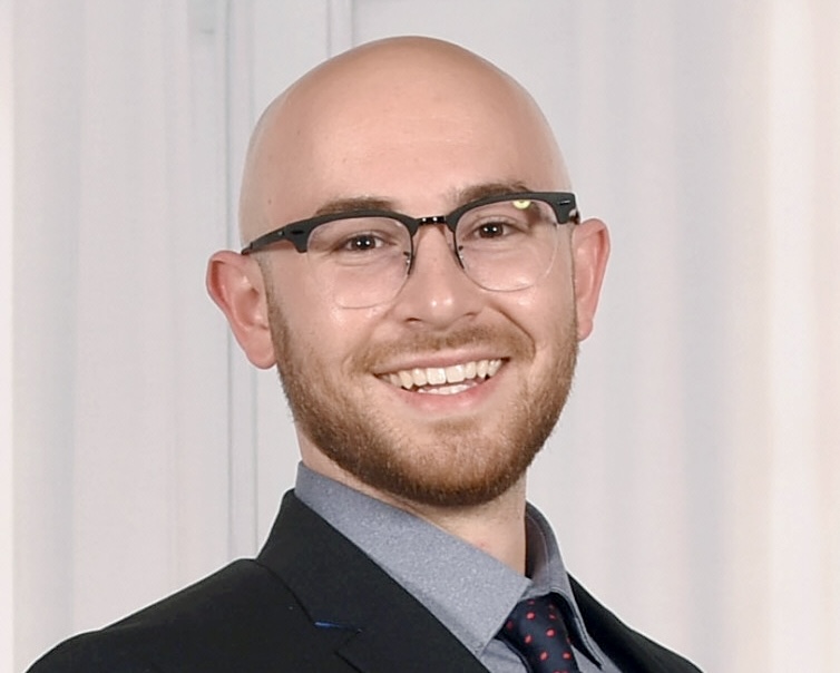 A smiling man with glasses and a beard, wearing a suit and tie, stands in front of a light-colored backdrop.