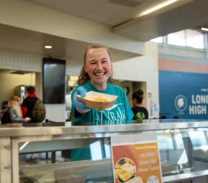 A smiling student in a turquoise shirt serves food at Longmont High School's cafeteria.