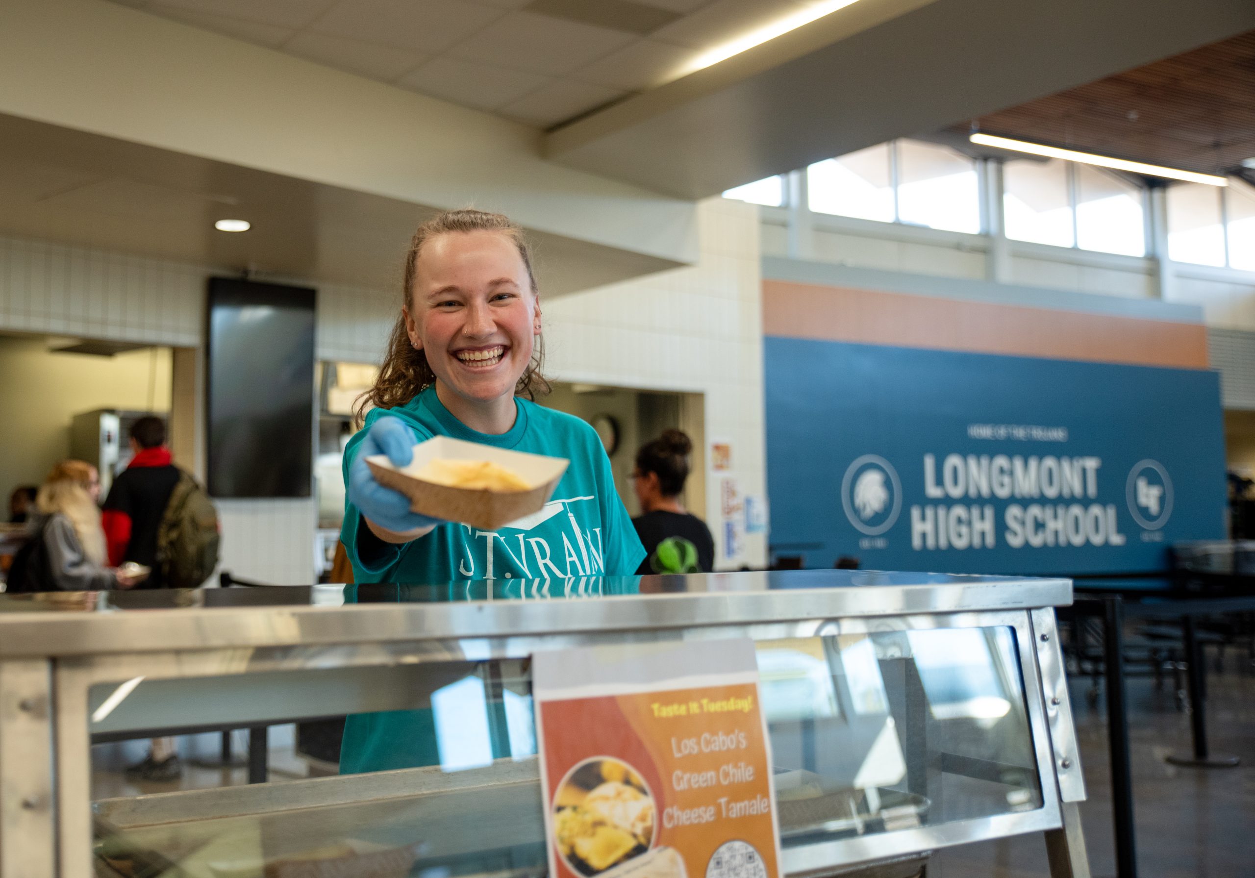 A smiling student in a cafeteria holds a tray of food, with a school sign in the background.