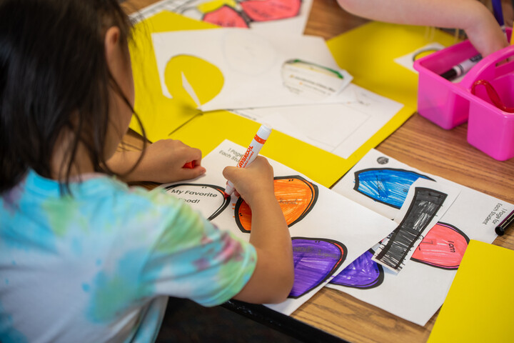 A child colors a paper project with markers on a table filled with colorful art supplies and papers.