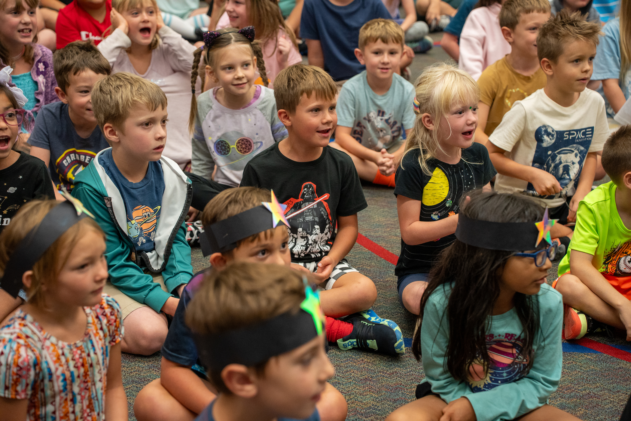 A group of children sitting on the floor, engaged and smiling, wearing colorful headbands with stars.